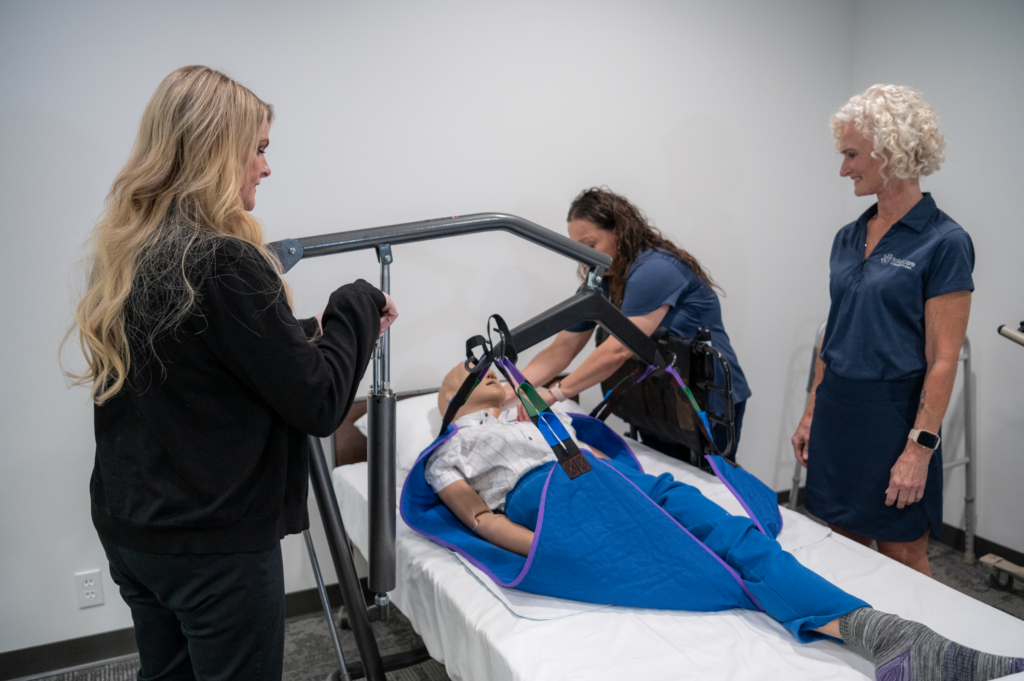 Caregiver receiving training on using patient lifting equipment with a mannequin on a bed, while paying attention to the trainer.